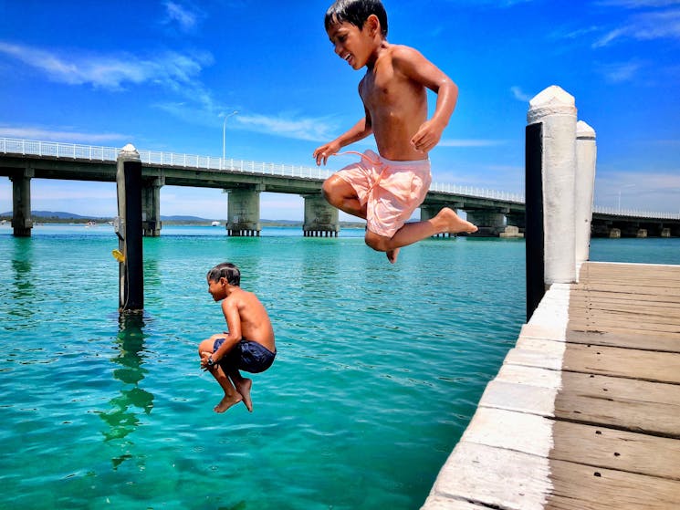 Two children jump joyfully into the clear waters of Wallis Lake, a scenic and family-friendly place to visit in NSW known for swimming and waterfront fun.