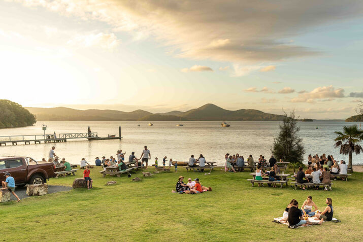 nsw-getaways NSW getaways by the water with people enjoying a grassy foreshore picnic area beside a calm lake, jetty and sunset sky