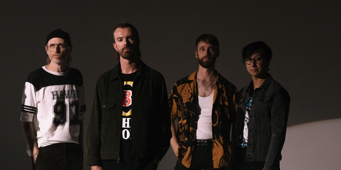 Four musicians standing together in a moody studio portrait, promoting a live music event that adds to the appeal of places to visit new south wales for relaxed coastal getaways.